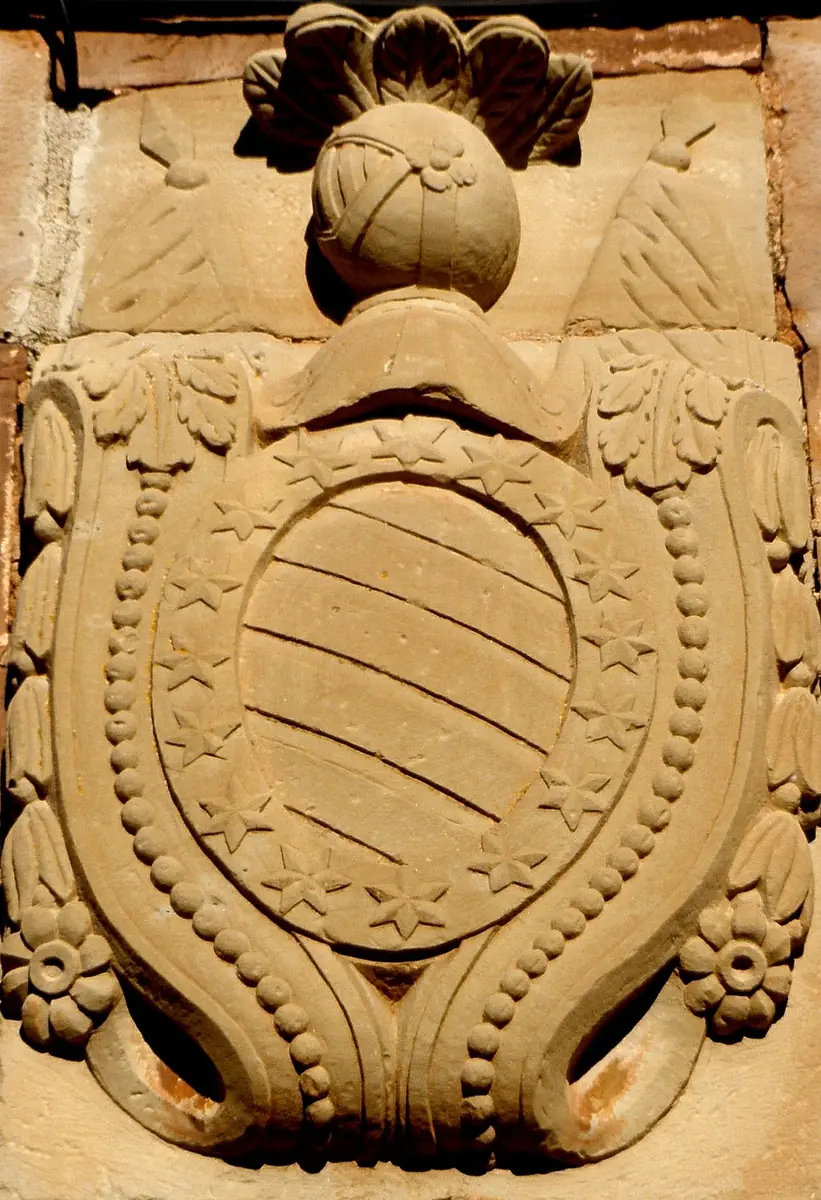 Escudo oval con bandas, orlado de estrellas y bajo un yelmo con penacho, en Sorlada.
