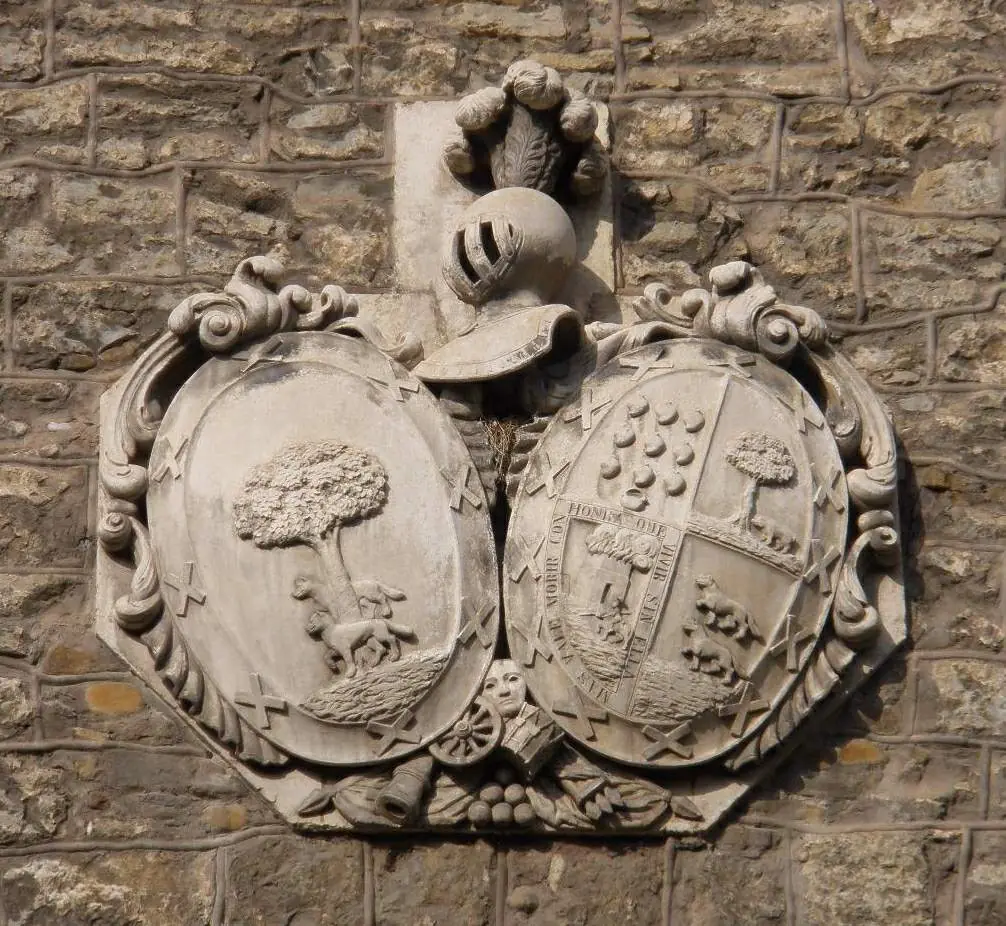 Dos escudos ovalados unidos bajo un mismo yelmo, uno con árbol y lobo y otro con varias figuras, con un lema en una cartela, en Bergara.