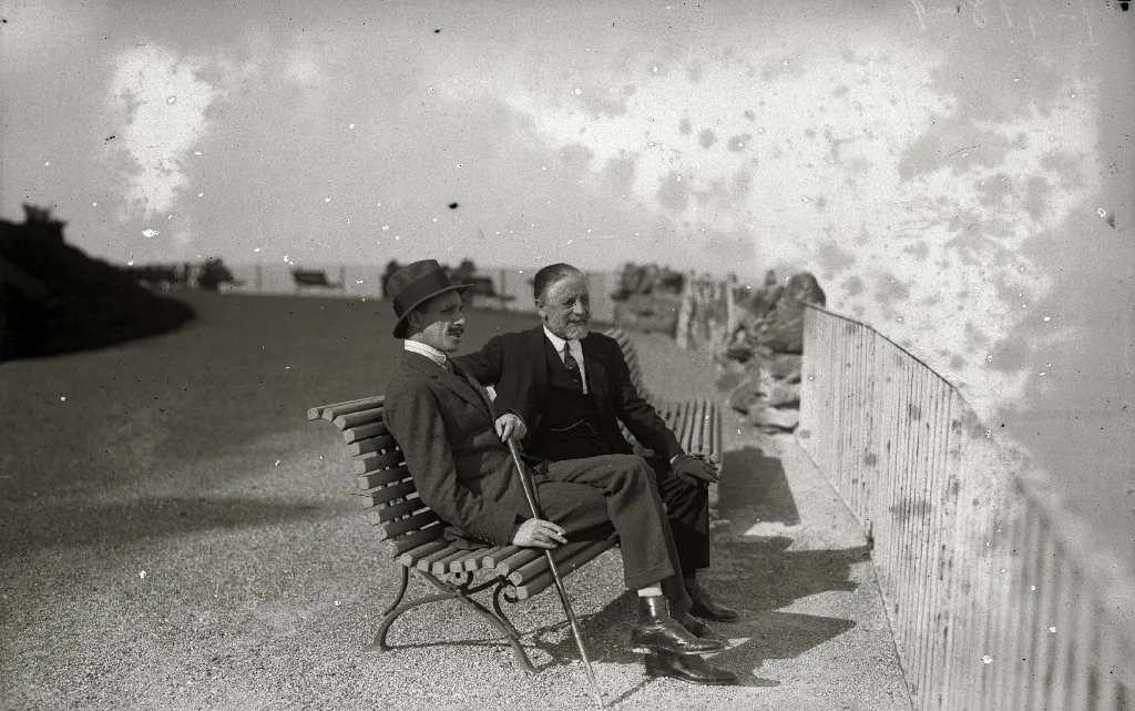 Dos hombres sentados en un banco del monte. Según el título del archivo, uno de ellos es el rey Alfonso XIII.