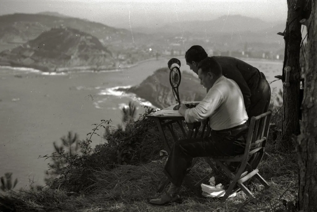 Un locutor ante un micrófono con la bahía de La Concha al fondo. Según el título del archivo, es José Zubimendi retransmitiendo las regatas para Unión Radio San Sebastián.