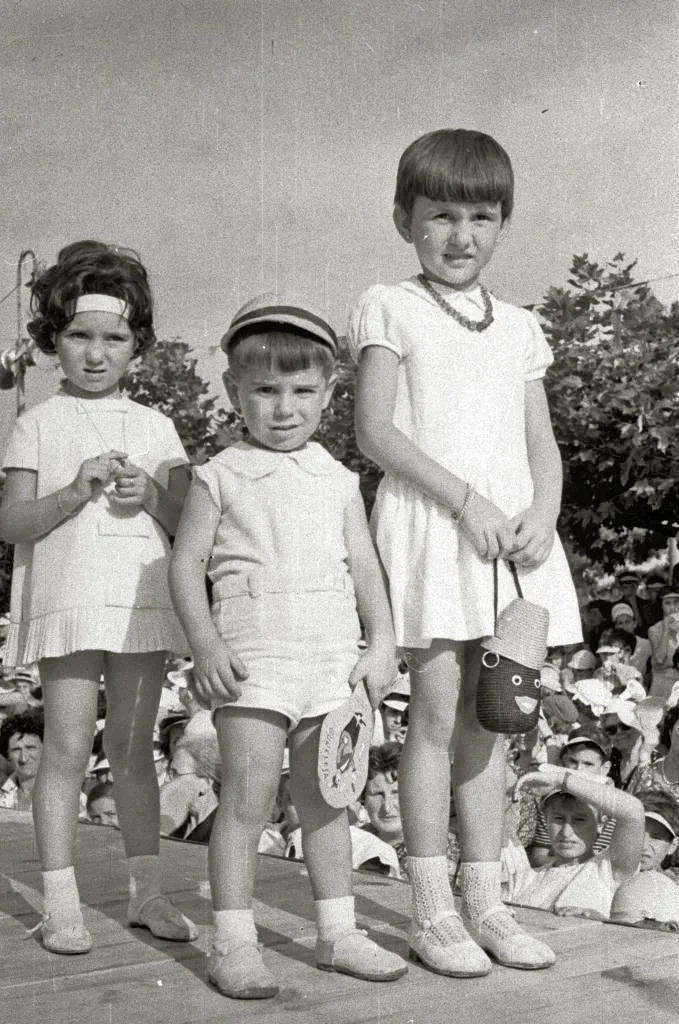 Tres niños sobre un entarimado con público detrás, en un desfile de moda infantil en el monte.